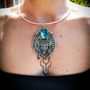 May include: A silver choker necklace with a large pendant featuring a detailed silver skull with a turquoise stone. The pendant is surrounded by smaller skulls and a sunburst design.