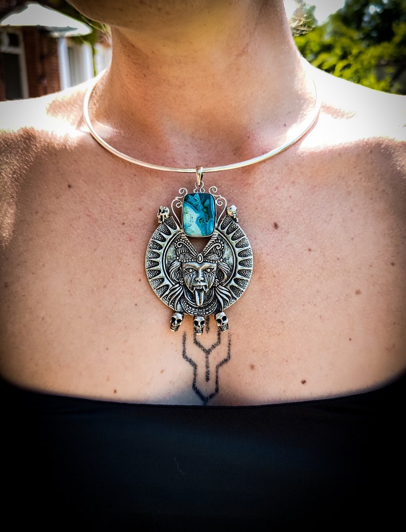 May include: A silver choker necklace with a large pendant featuring a detailed silver skull with a turquoise stone. The pendant is surrounded by smaller skulls and a sunburst design.