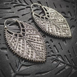 Peut inclure: Une paire de boucles d'oreilles argentées avec un design géométrique en forme de feuille. Les boucles d'oreilles sont grandes et ont une forme pendante.