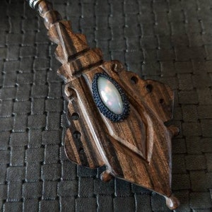 Peut inclure: Un pendentif en bois avec des sculptures complexes et une pierre précieuse d'opale blanche sertie dans une lunette en métal noir. Le pendentif est attaché à un cordon de perles noires.