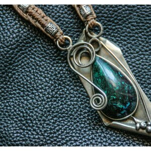 Peut inclure: Un collier avec un pendentif en argent orné d'une pierre précieuse verte et de fils métalliques travaillés. Le pendentif a la forme d'une larme et est suspendu à un cordon brun.