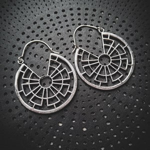 Peut inclure: Une paire de boucles d'oreilles créoles en argent avec un motif géométrique. Les boucles d'oreilles sont en métal et ont une forme circulaire. Le motif est complexe et présente une série de lignes et de courbes.