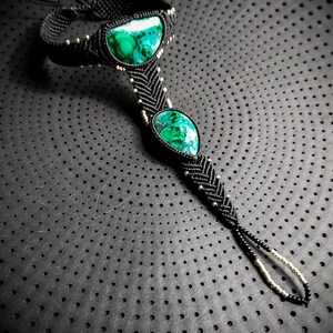 May include: A black macrame ankle bracelet with three green malachite stones. The bracelet is on a black background with a dotted pattern.