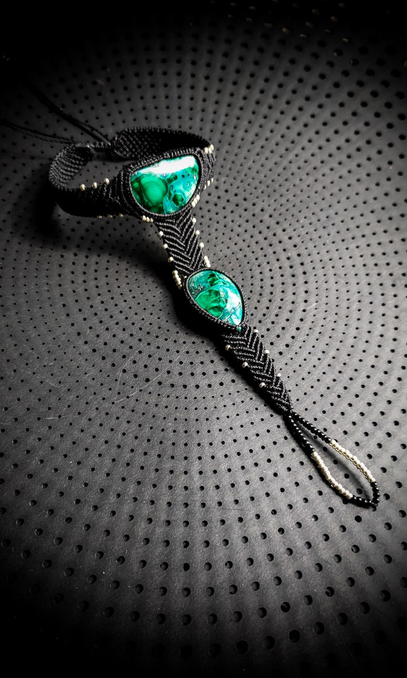 May include: A black macrame ankle bracelet with three green malachite stones. The bracelet is on a black background with a dotted pattern.