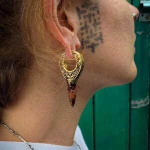 May include: A gold and brown ear gauge with an intricate design. The gauge is worn in the left ear of a person with short blonde hair.
