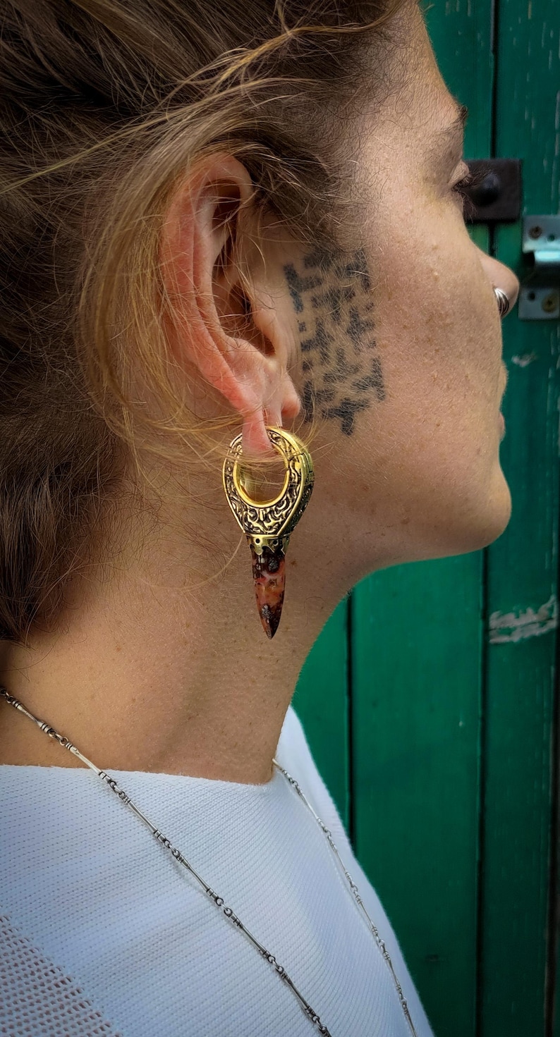 May include: A gold and brown ear gauge with an intricate design. The gauge is worn in the left ear of a person with short blonde hair.