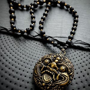 May include: A black lava rock beaded necklace with a gold-toned pendant featuring a detailed, ornate, and slightly menacing face.