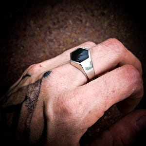 Peut inclure: Bague en argent avec une pierre noire hexagonale. La bague présente un design simple et moderne. La bague est portée sur une main tatouée.