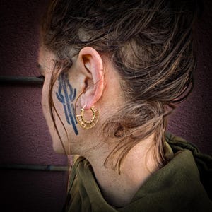 May include: A person wearing a gold hoop earring with a geometric design. The earring is in the shape of a circle with a series of small, evenly spaced lines radiating outward from the center.