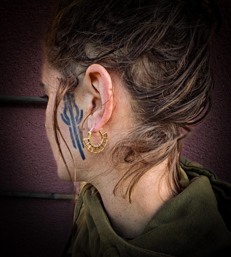 May include: A person wearing a gold hoop earring with a geometric design. The earring is in the shape of a circle with a series of small, evenly spaced lines radiating outward from the center.