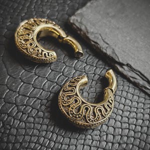 Op de afbeelding: Twee gouden metalen oorpluggen met ingewikkelde, sierlijke ontwerpen. De pluggen liggen op een zwart, getextureerd oppervlak.