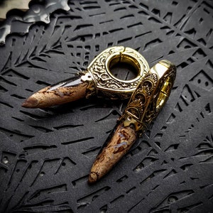 May include: A pair of gold-toned ear gauges with intricate designs and brown and black stone accents. The gauges are on a black textured surface.
