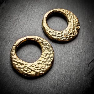 Può includere: Due orecchini a cerchio in metallo color oro con una texture martellata. Gli orecchini sono circolari e hanno una finitura liscia e lucida.