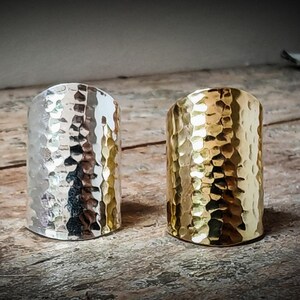 Pode incluir: Dois anéis de metal martelado, um prateado e outro dourado, sobre uma superfície de madeira.