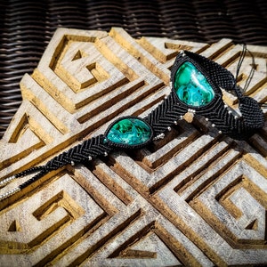 May include: Black macrame bracelet with two green malachite stones. The bracelet is on a wooden surface with a geometric pattern.