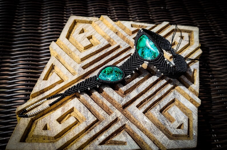 May include: Black macrame bracelet with two green malachite stones. The bracelet is on a wooden surface with a geometric pattern.