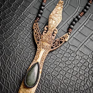 Peut inclure: Un collier pendentif artisanal avec un pendentif sculpté de couleur os, orné de motifs complexes et d'une pierre verte centrale. La chaîne du collier est composée de perles noires, posé sur une surface noire texturée.