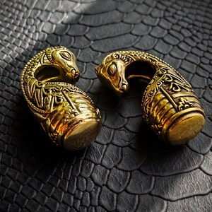 Puede incluir: Dos expansores dorados ornamentados con un dise&ntilde;o de serpiente. Los expansores est&aacute;n detallados con patrones intrincados y se asientan sobre una superficie oscura texturizada. Las cabezas de serpiente se curvan una hacia la otra.