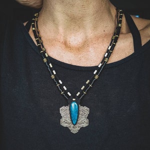 Op de afbeelding: Een zwarte koordketting met een geometrische hanger met een turquoise steen. De hanger is gemaakt van messing en heeft een gedetailleerd ontwerp.