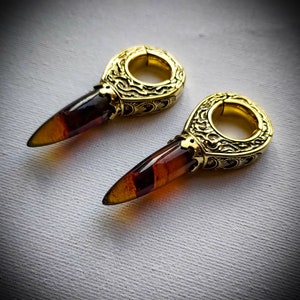 May include: A pair of gold-toned earrings with a detailed design and a pointed amber stone. The earrings are designed to be worn in stretched earlobes.