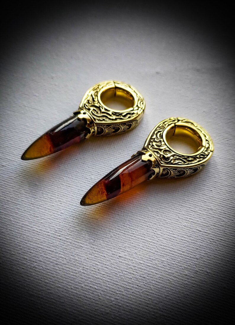 May include: A pair of gold-toned earrings with a detailed design and a pointed amber stone. The earrings are designed to be worn in stretched earlobes.