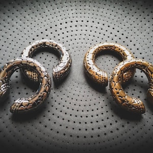 Peut inclure: Deux paires d'écarteurs d'oreilles décoratifs, une paire est argentée et l'autre dorée. Les deux paires ont un design texturé et écailleux.