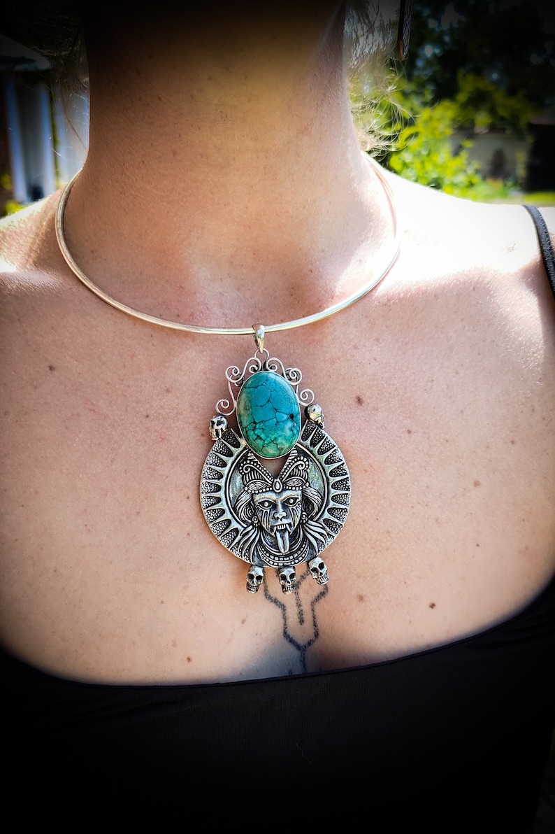May include: A silver choker necklace with a pendant featuring a turquoise stone and a detailed silver design of a face with a long tongue. The pendant is surrounded by a circle of skulls.