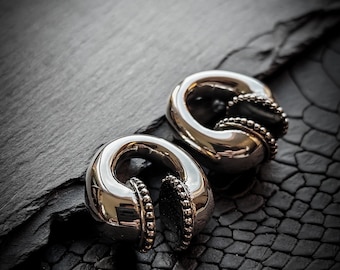 May include: Two silver ear gauges with a beaded edge. The gauges are on a black textured surface.