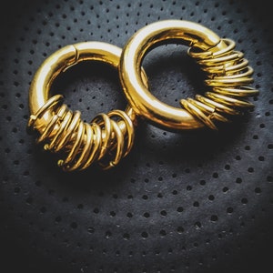 May include: Two gold hoop earrings, one with a stack of smaller hoops, on a black textured background.
