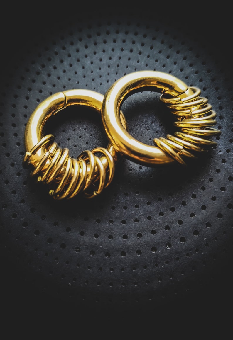 May include: Two gold hoop earrings, one with a stack of smaller hoops, on a black textured background.