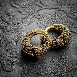 May include: Two ornate, gold-colored earrings with intricate, swirling patterns. The earrings are circular and have a detailed, textured surface. They rest on a dark, textured background with a floral design, creating a striking contrast.