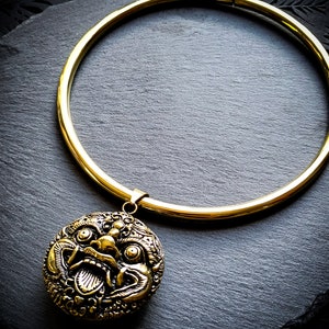 May include: A gold choker necklace with a large, detailed, gold pendant featuring a mythical creature's face. The necklace is on a black background.