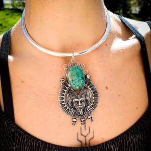 May include: A silver choker necklace with a large turquoise stone pendant. The pendant features a detailed design of a face with a long tongue and skulls.