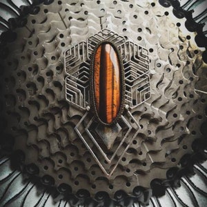 May include: A silver geometric pendant with a tiger's eye stone set in the centre. The pendant is displayed on a background of stacked metal gears.