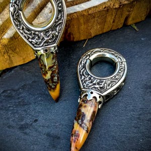 May include: Two silver and gold ornate ear gauges with a pointed, multi-colored stone tip. The gauges are on a dark gray surface.