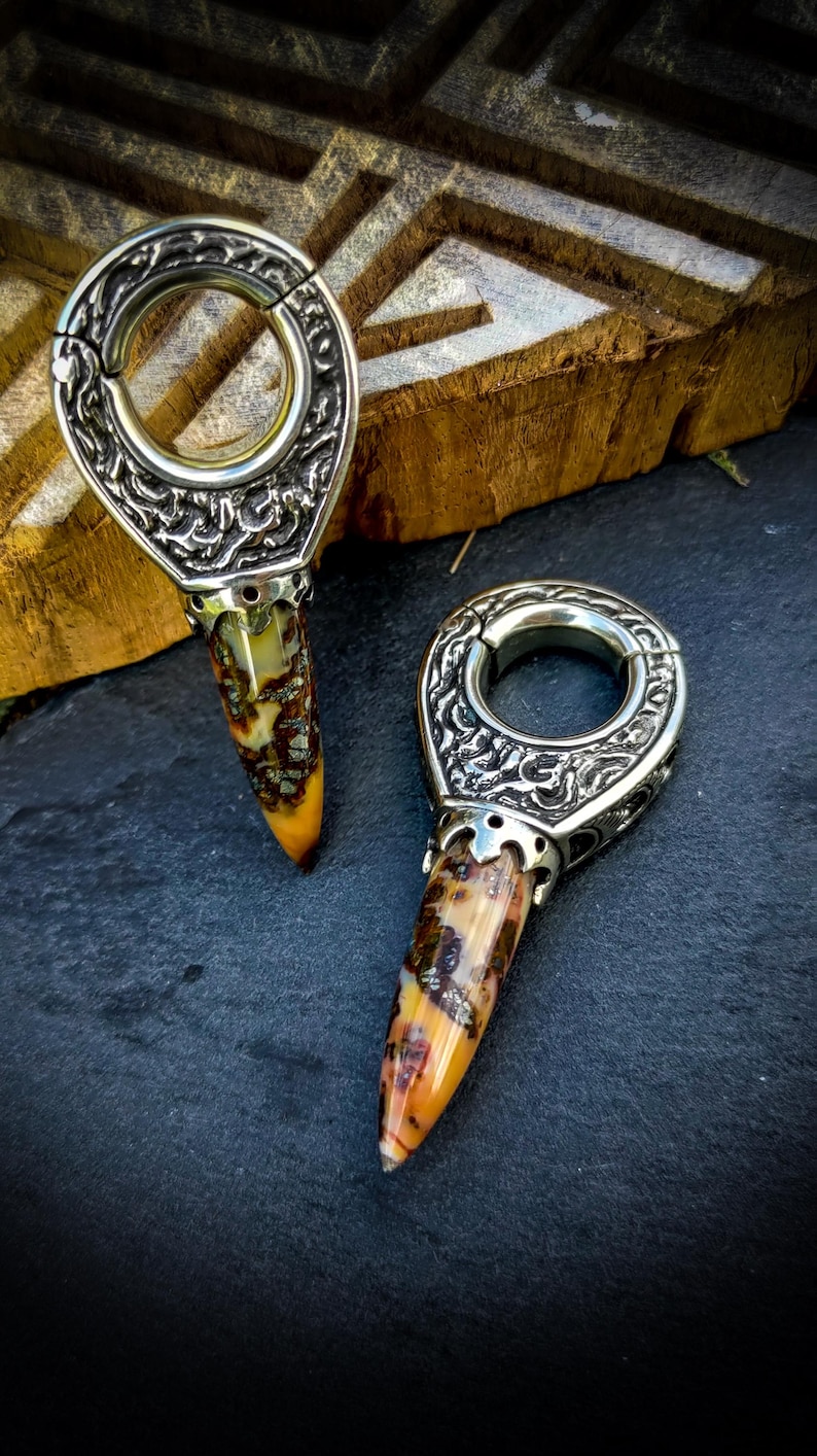 May include: Two silver and gold ornate ear gauges with a pointed, multi-colored stone tip. The gauges are on a dark gray surface.