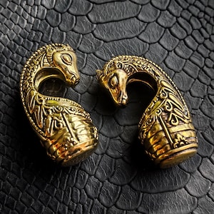 Puede incluir: Dos expansores de oreja dorados y ornamentados con un dise&ntilde;o de cabeza de caballo. Los expansores presentan patrones intrincados y se encuentran sobre una superficie negra texturizada. Las cabezas de caballo se curvan hacia adentro, creando una forma &uacute;nica.
