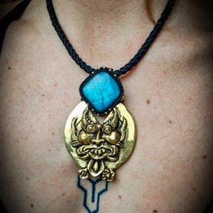 Peut inclure: Un collier unique avec un pendentif doré orné d'un visage élaboré, associé à une pierre précieuse carrée bleu clair. Le pendentif est suspendu à un cordon tressé noir. Un bijou remarquable.
