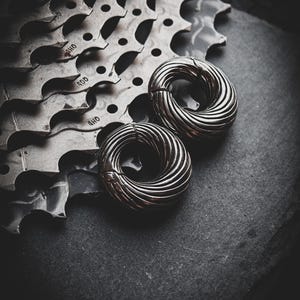 Puede incluir: Dos anillos de metal plateado con un diseño estriado, que descansan sobre una superficie gris oscuro. Los anillos son de tamaño y forma similares, con una abertura circular en el centro.