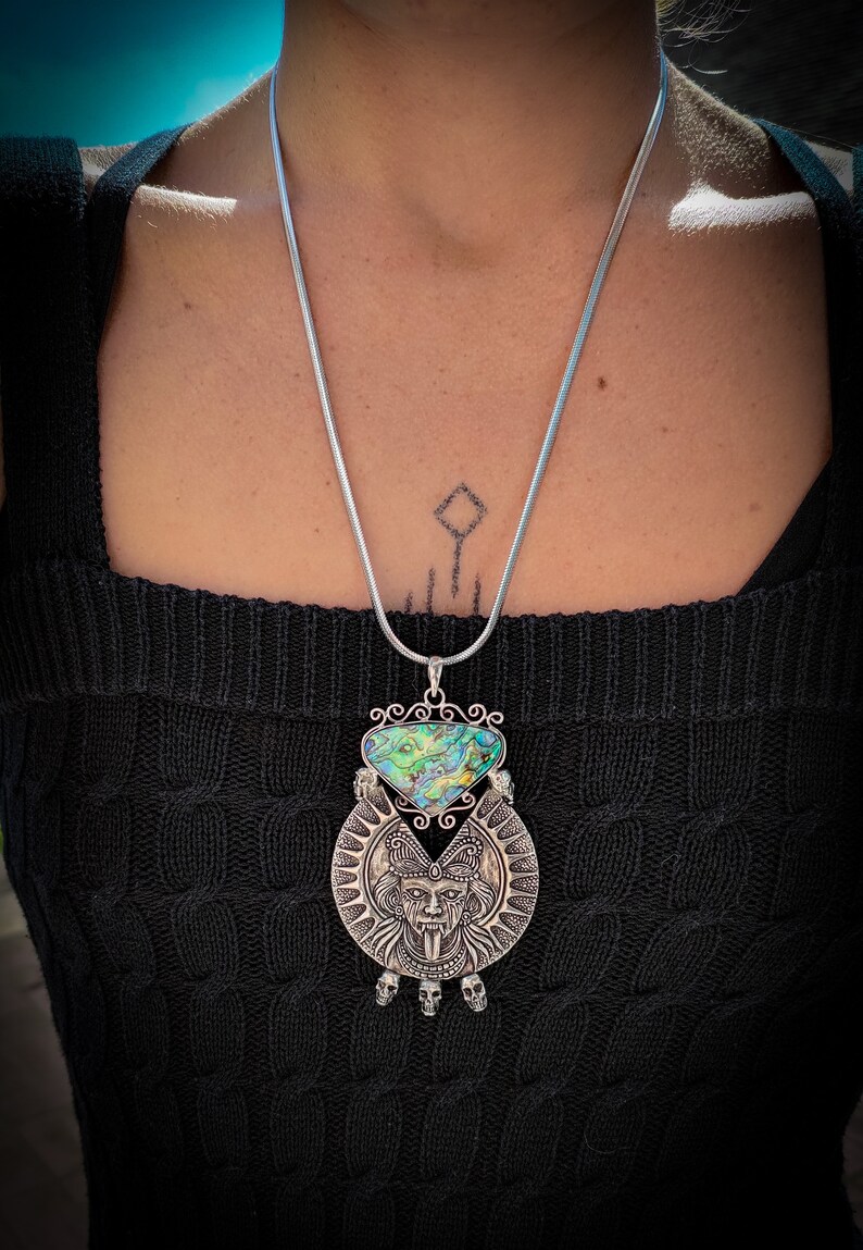 May include: A silver chain necklace with a large pendant featuring a green and blue abalone shell and a silver skull design.