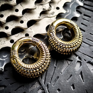 May include: Two gold-colored metal ear gauges with intricate designs. The gauges are on a black textured surface.
