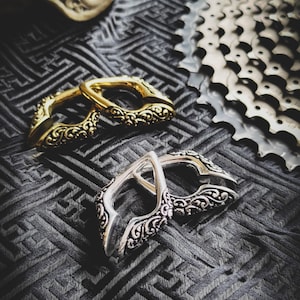 Peut inclure: Deux boucles de ceinture en métal ornées, l'une dorée et l'autre argentée, avec des motifs floraux complexes. Les boucles sont posées sur un tissu noir à motif géométrique.