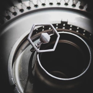 Peut inclure: Une bague hexagonale argentée avec un motif hexagonal plus petit au centre. La bague est posée sur une surface en métal argenté et noir.