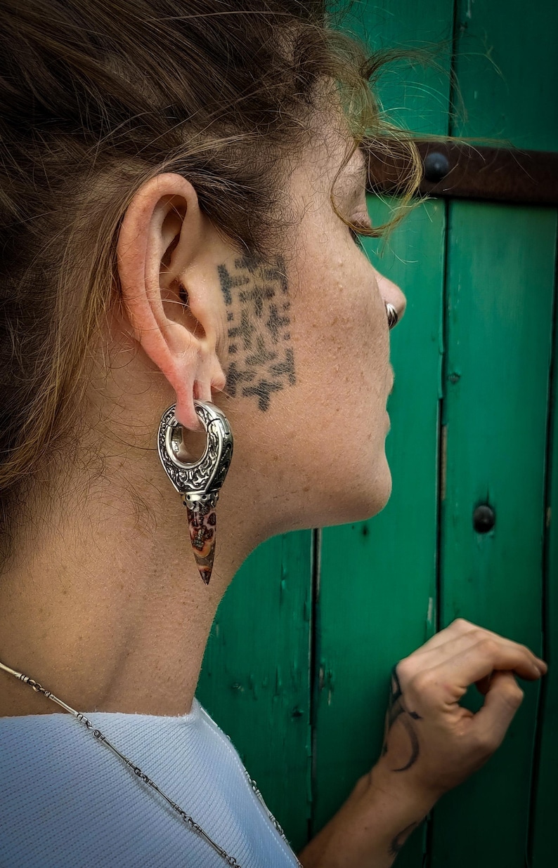 May include: A person with a black and white tattoo on their face is wearing a silver and brown ear gauge with a pointed tip. The gauge is a large hoop with intricate designs.