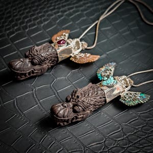 Peut inclure: Deux colliers pendentifs dragon. Les têtes de dragon sont sculptées dans un matériau brun foncé, avec des accents argentés et des ailes. Un pendentif a une gemme rouge, l'autre a des ailes turquoise et irisées. Les colliers reposent sur une surface noire texturée.