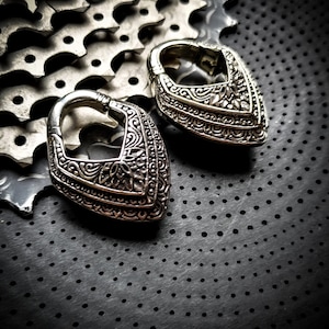Può includere: Due gauge per orecchie in argento ornate con un design floreale dettagliato. Le gauge hanno la forma di un cuore con una parte superiore arrotondata e una parte inferiore appuntita.