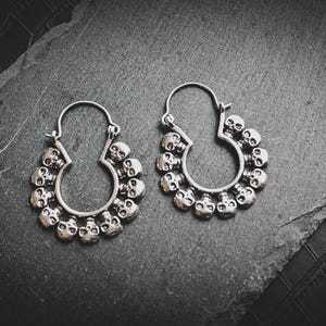 Peut inclure: Une paire de boucles d'oreilles créoles en argent avec un motif de crâne. Chaque boucle d'oreille a un cercle de crânes autour du cercle.