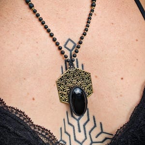 Peut inclure: Collier de perles noires avec un pendentif géométrique doré et une pierre ovale noire. Le collier est porté sur la peau et un haut en dentelle noire.