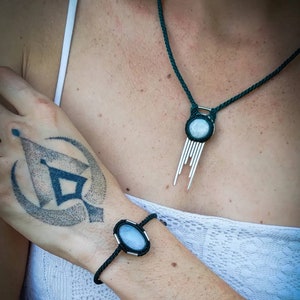Puede incluir: Un collar de cordón negro con un colgante de plata que presenta una piedra redonda de color azul claro y una franja de hilos plateados. Una pulsera de cordón negro a juego con una piedra azul claro similar.