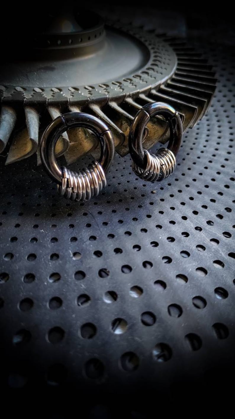 May include: Two black circular rings with silver metal rings stacked on top of each other. The rings are on a black surface with a pattern of small holes.